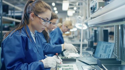 Wall Mural - Female Electronics Factory Worker in Blue Work Coat and Protective Glasses is Assembling Laptop's Motherboard with Tweezers and Screwdriver. High Tech Factory Facility with Multiple Employees. 