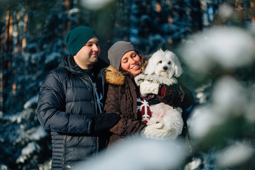 Cute young couple having fun in winter forest with their pretty little white dog. Man and woman in sweater with a snowflake. Christmas and winter holidays