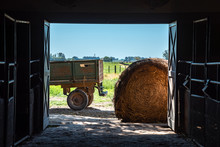 Inside A Barn Looking Outside, With A Hay Roll Or Bale And A Truck Trailer With A Blue Sky And Green Grass
