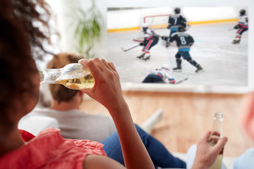 sport, entertainment and people concept - woman with friends drinking beer and watching ice hockey game on projector screen at home