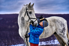 Young Pretty Woman In A Blue Jacket And A Sports Hat For A Walk With A White Horse In The Winter Cloudy Day.