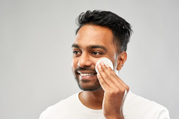 grooming, skin care and people concept - smiling young indian man cleaning his face with cotton pad over grey background