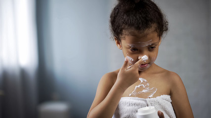 Funny little girl putting cream on nose, wearing bathrobe and copying mom