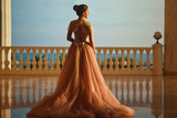 Beautiful woman in luxurious ballroom dress with tulle skirt and lacy top standing on the large balcony with sea view. Back view