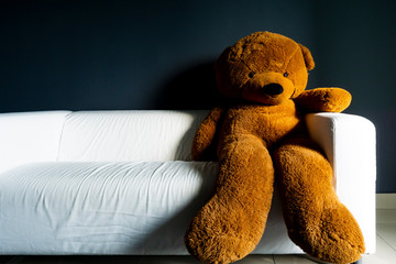 Brown color teddy bear sitting on a white sofa with grey background