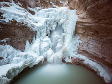 A Half Frozen Waterfall Flows Into A Green Pool Surrounded By Huge Ice Masses From A Cold Winter