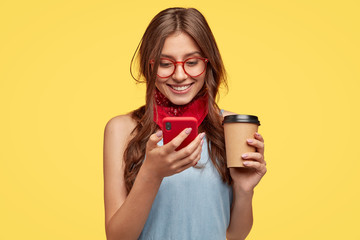 Cheerful girl has coffee break, rejoices buying new gadget, reads notification on red mobile phone, updates favourite app, types message and smiles at screen, wears glasses, isolated over yellow wall