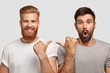 © WHstudio Leushin N - Shocked unshaven man with dark stubble indicates at his companion, wears grey t shirt. Cheerful ginger guy with trendy haircut and bristle points at fellow. Two friends model indoor over white wall