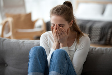 Upset young female sit on couch at home crying over ended relationships, unhappy girl cover face feeling down after breaking up with boyfriend, woman lie on sofa have problems in life