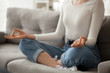 © fizkes - Close up of focused female sit on cozy couch meditating with mudra hands, calm woman practice yoga in lotus position on sofa at home, girl manage stress controlling emotions. Healthy lifestyle concept