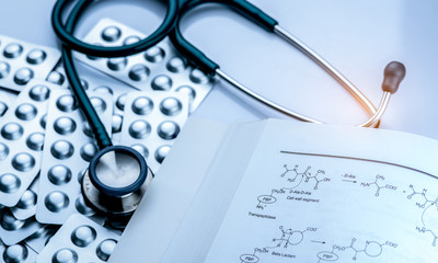 Pile of tablets pill in blister pack with drug structure book and stethoscope on white background. Medical equipment for diagnosis. Pharmacology textbook. Drug research concept. Expert in medicine.