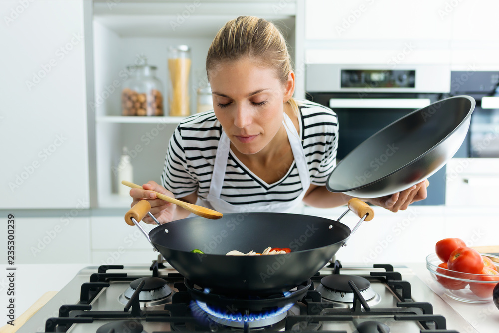 Healthy young woman cooking and smelling food in frying pan in the kitchen at home. - obrazy, fototapety, plakaty 
