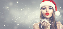 Christmas Winter Fashion Girl On Holiday Blurred Winter Background. Beautiful New Year And Xmas Holiday Makeup. Beauty Model Woman In Santa's Hat Blowing Snow In Her Hand