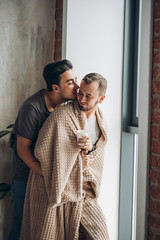 Wall Mural - Handsome brunette gay guy hugging his romantic friend, covered with plaid, while standing at the window of mofern loft hotel room. Non-traditional orientation, closeness, tenderness concept.