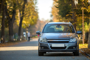 Wall Mural - Front view of gray shiny empty car parked in quiet area on asphalt road at lamppost on blurred green and yellow trees bokeh background on bright sunny day. Transportation and parking concept.