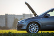 © bilanol - Side view detail of car with open hood on empty gravel field road on blurred apartment building and clear bright sky copy space background. Transportation, vehicles problems and breakdowns concept.