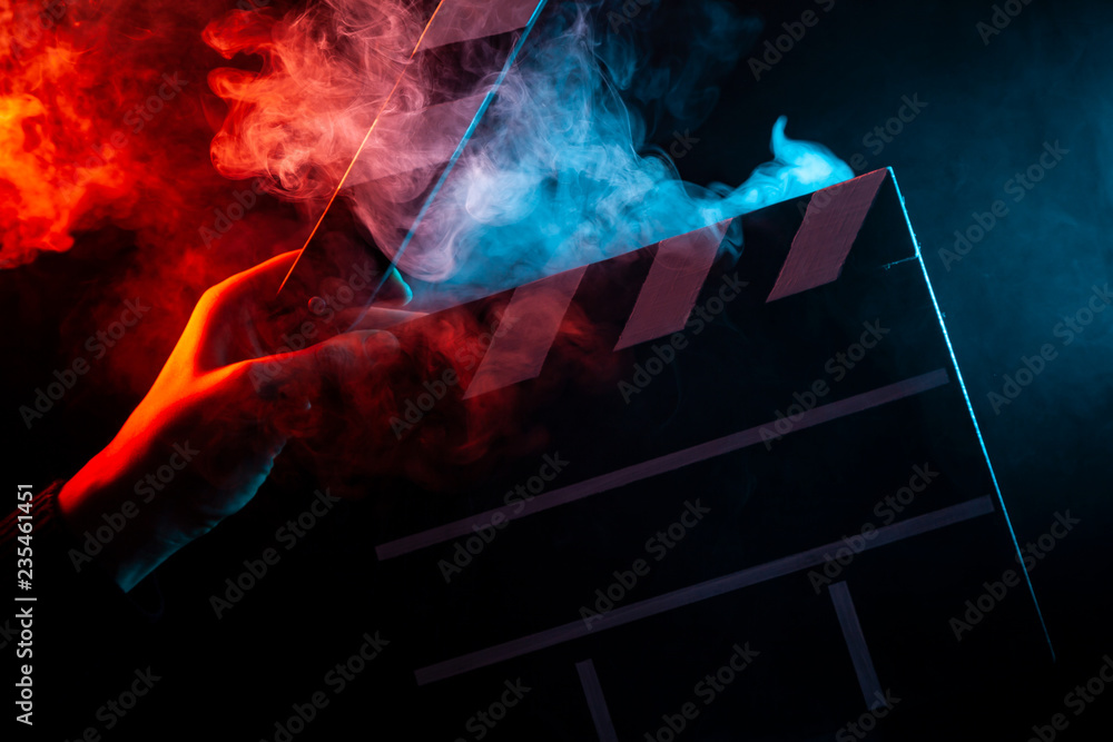 Close-up on an open clapper in hand before starting shooting a film with multi-colored smoke around with red and blue backlighting on a black isolated background - obrazy, fototapety, plakaty 