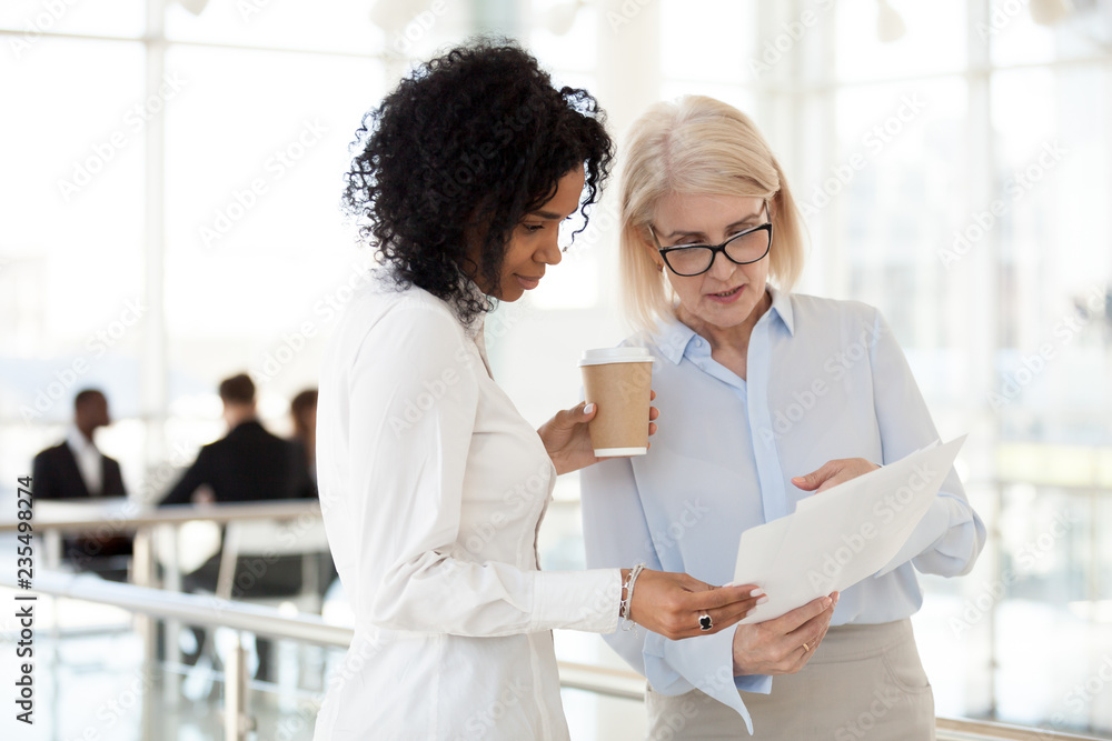 Senior mature caucasian female executive ceo discuss report with young mixed race black colleague, serious old aged mentor reads paper explains business plan to african employee intern talk in office