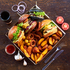 Wall Mural - On a wooden background, 4 different burgers, various hot fried foods: potatoes, balls and deep-fried rings. around the glass with a drink, sliced tomatoes, cutlery, onion rings, red sauce and garlic.