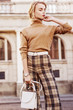 Outdoor fashion portrait of young beautiful fashionable girl wearing trendy beige cashmere turtleneck, high-waisted checkered trousers, belt, wrist watch, holding small white bag, posing in street