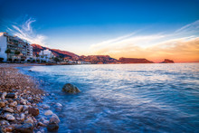 Long Exposure Of The Rocky Shore And Blue Water Of Altea Spain With Buldings And Mountains In The Distance And The Orange Painted Sky From The Setting Sun Mixed With Blue