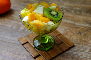 Cup of fresh fruit salad with different tropical fruit on a wooden table with raw fruits in the background. the ingredients are kiwi, persimmon, pineapple and apple