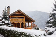 Christmas Wooden Mansion In Mountains On Snowfall Winter Day. Cozy Chalet On Ski Resort Near Pine Forest. Cottage Of Round Timber With Wooden Balcony. Fir-trees Covered With Snow. Chimneys Of Stone.