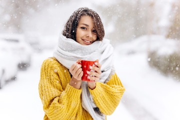 Smiling dark-haired girl in a yellow sweater, jeans and a white scarf standing with a red mug on a snowy street on a winter day