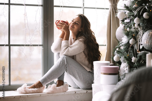 Fotografía Nice dark-haired girl dressed in pants, sweater and warm slippers holds a red cu
