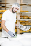 Fototapeta Do przedpokoju - Man rubing cheese wheels with wax at the cheese manufacturing with shelves full of cheese on the background