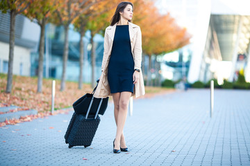 Girl carrying suitcase by streets. Smiling blond businesswoman with wheeled luggage