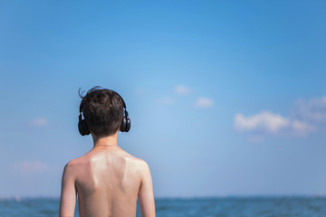 Wall Mural - Happy cheerful teen boy on the beach with the headphones running on the wave of the sea on the resort town background. Concept