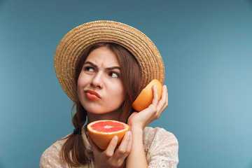 Sticker - Confused young beautiful woman isolated over blue wall holding citrus near ear wearing hat.