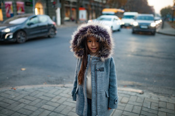 Child girl is standing at the sidewalk on background of city street in evening.