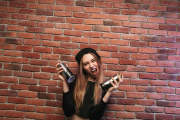 Image of subcultural hip hop girl 20s, standing against brick wall with spray cans