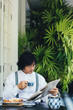 © LUMINA IMAGES - Serious middle-aged Asian businessman having a breakfast at hotel terrace and reading newspaper while drinking a tea.