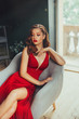© kharchenkoirina - Hot young adult and imperious woman, dressed in a long scarlet red long dress, sexually shows her nude elegant leg, sitting on a grey chair in a loft room with window to the floor, daring style