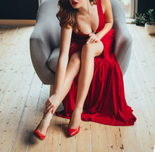 Sexy Red-haired Young Woman Playfully Touches Her Fingers Over Her Bare Leg, Showing The Neckline. In A Long Scarlet Expensive Delightful Long Luxurious Dress And Red High-heeled Shoes