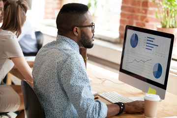 Wall Mural - Serious African American man in glasses talking by phone about economic report with pie diagrams, marketing plan, business results, using computer, applications, looking at screen