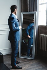 Wall Mural - Perfect look. Reflection of handsome young man in full suit adjusting his jacket while standing in front of the mirror indoors.