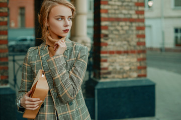 Wall Mural - Outdoor portrait of young beautiful fashionable lady wearing stylish checkered blazer, beige cashmere turtleneck, holding small suede bag, posing in street of european city. Copy, empty space for text