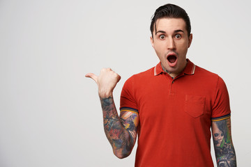 Stylish man fascinated and excited. Close up of amazed attractive young male in red polo pointing aside with dropped jaw, standing over white wall