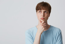 Thoughtful Focused European Young Man In Blue T-shirt, Keeps Arm On Chin And Looking Distance, Furrowed Eyebrows, Being Confused And Questioned, Thinking What Do To Have Fun, Troubled, Over White Wall