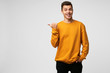 Young attractive handsome guy looks delighted gladden amazed, mouth open, smiling, like dream came true, big discount in shop,wears orange sweatshirt, points with thumb finger on a blank copyspace