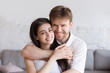 © fizkes - Head shot portrait of loving embracing happy couple in living room, smiling boyfriend standing behind attractive girlfriend, hugging, man and woman holding hands, family portrait, look at camera