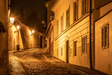 Fototapeta Uliczki - Narrow cobbled street in old medieval town with illuminated houses by vintage street lamps, Novy svet, Prague, Czech Republic. Night shot.