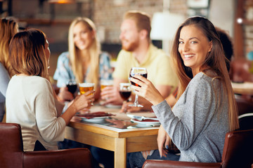 Woman looking at camera and holding glass of wine while sitting in restaurant. In background friends drinking and chatting.