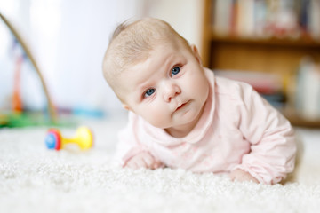 Wall Mural - Adorable newborn baby girl lying on belly in white sunny bedroom or nursery. Textile and bedding for children. Cute girl with blue eyes at home playing with colorful rattle toys.