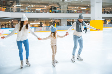 Wall Mural - family in sweaters holding hands while skating together on ice rink