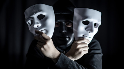 Mystery hoody man wearing black mask holding two white masks in his hand. Anonymous social masking. Major depressive disorder or bipolar disorder. Halloween concept